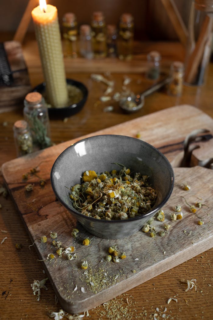 Rustic wooden scene with dried herbs in a ceramic bowl surrounded by candles and bottles.
